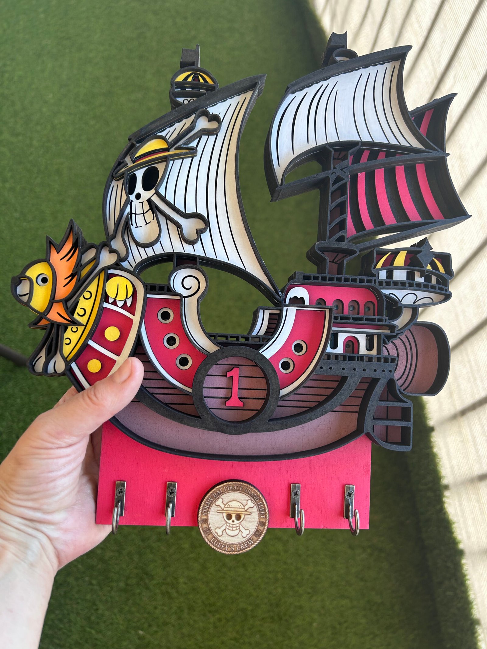 Colgador de llaves artesanal de madera 3D del barco One Piece Thousand Sunny, mostrando el modelo detallado en capas y los cuatro ganchos metálicos para llaves.